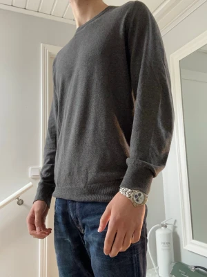 Mörkgrå tröja Dressmann - En enkel och stilren mörkgrå tröja med rund hals och långa ärmar. Tröjan har ribbade muddar vid ärmslut och nederkant. Perfekt att matcha med jeans för en clean look.