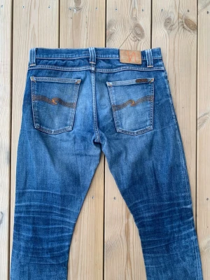 Nudie Jeans Tube Tom - Säljer ett par klassiska blå jeans från Nudie Jeans med snygg slitning och kontrastsömmar. Jeansen har raka ben, två bakfickor med vågig brodyr och patch i läder bak i midjan. Perfekta för en avslappnad och trendig look.