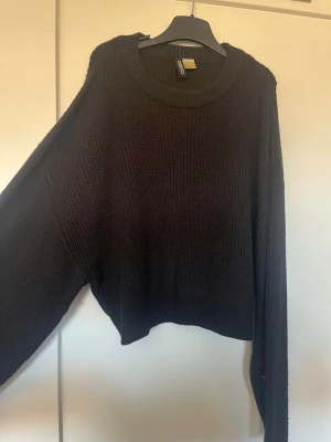 Svart stickad tröja från H&M Divided - Mysig svart stickad tröja från H&M Divided med rund halsringning och avslappnad passform. Tröjan har långa ärmar och ribbade muddar vid hals, ärmslut och nederkant. Perfekt för lager på lager och enkel att styla till olika outfits.