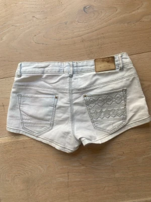 Ljusblå denimshorts - Säljer ett par vintage ljusblå denimshorts med klassisk femficksdesign och snygga broderade detaljer på ena bakfickan. Shortsen har låg midja och är tillverkade i bomull med en liten mängd stretch för extra komfort.