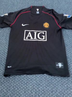 Manchester United Ronaldo tröja svart - Snygg svart Manchester United matchtröja från Nike med röda detaljer och AIG som sponsor. Tröjan har klubbmärke på bröstet, nummer 7 och Ronaldo på ryggen. Tillverkad i lätt och ventilerande material, perfekt för fotboll eller träning. (OBS)  Pris kan sänkas rätt så mycket vid äffar!