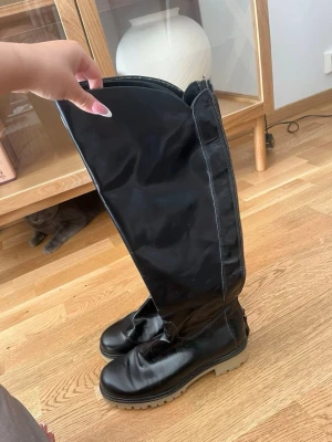 Svarta höga stövlar med grov sula - Säljer ett par svarta höga stövlar med dragkedja längs skaftet och grov beige sula. Stövlarna är i glansigt material, troligen konstläder, och har en rymlig insida. Perfekta för dig som vill ha ett statement-plagg till din outfit.