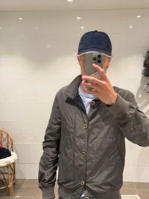 Aspesi bomberjacka med knappar - Snygg olivgrön bomberjacka med ribbade muddar och krage. Jackan har stora knappar framtill och en avslappnad passform. Tillverkad i ett lätt, glansigt material som ger en modern look. Perfekt för dig som vill ha en stilren och enkel jacka till vardags.