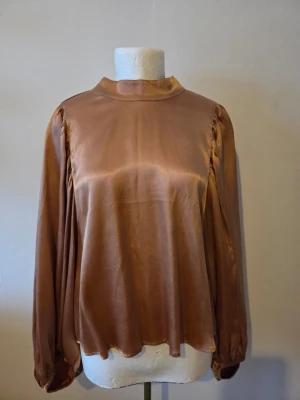 H&M satinblus med hög krage - Snygg kopparfärgad blus i glansigt satinmaterial med hög krage och långa, lösa ärmar. Blusen har en avslappnad passform och subtila rynkdetaljer vid axlarna. Perfekt för dig som gillar en stilren och trendig look.