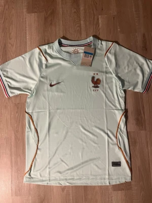 Frankrike VM-2026 Fotbollströja  - Snygg ljusgrön fotbollströja med Frankrikes landslagsmärke och två stjärnor på bröstet. Tröjan har korta ärmar med randiga muddar och orange detaljer längs sidorna. Perfekt för match eller träning. HELT NY
