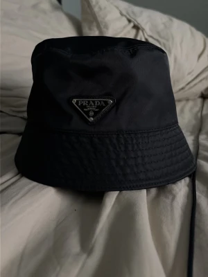 Svart bucket hat från Prada - Snygg svart bucket hat från Prada med det klassiska triangulära metallogot på framsidan. Hatten är tillverkad i nylon och har en mjuk, rundad form som ger en avslappnad vibe. Perfekt accessoar för att lyfta din streetwear-look.
