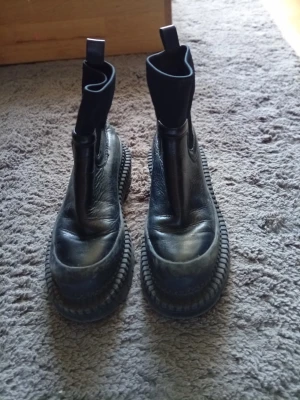 Chunky svarta Chelsea boots - Säljer ett par chunky svarta Chelsea boots med höga skaft och breda, grova sulor. Skorna har elastiska paneler på sidorna och dragflikar upptill för enkel påtagning. Tillverkade i svart läder med cool, robust look.