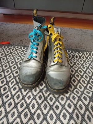 Silvriga Dr. Martens stövletter - Unika silvriga Dr. Martens stövletter med kontrasterande skosnören i blått och gult. Klassisk hög modell med grov sula och tydliga sömmar. Tillverkade i läder och har den ikoniska Dr. Martens-dragfliken baktill. Perfekt för dig som vill sticka ut.