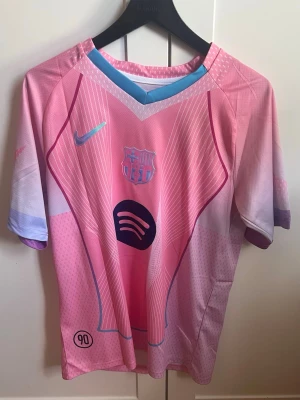 Rosa FC Barcelona matchtröja Nike - Snygg rosa FC Barcelona matchtröja från Nike med klubbmärke och Spotify-logga på bröstet. Tröjan har korta ärmar, V-ringning med blå detaljer och ett sportigt mönster i olika nyanser av rosa och lila. Perfekt för dig som älskar fotboll och vill sticka ut på planen.