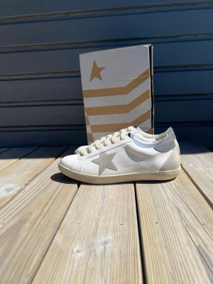 Golden Goose vita sneakers med stjärna - Snygga vita sneakers från Golden Goose med grå stjärndetalj på sidan och grå hälflik. Skorna har beigea skosnören och en klassisk låg siluett. Tillverkade i läder med slitstark gummisula. Perfekta för en clean och trendig look.