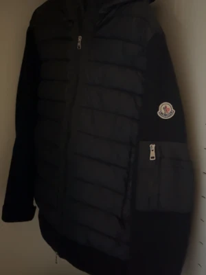 Moncler cardigan  - Säljer en svart dunjacka från Moncler med huva och dragkedja framtill. Jackan har Moncler-logga på ärmen och en praktisk ficka med dragkedja på ena ärmen. Klassisk quiltad design och sportig look, perfekt för kalla dagar.