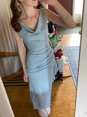 Ljusblå långklänning med slits - Säljer en elegant ljusblå långklänning med draperad urringning och hög slits på sidan. Klänningen har figurnära passform och fina detaljer i midjan. Klänningen är helt ny och inte använd mer än att jag testat den. 