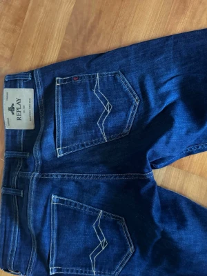 Mörkblå jeans från Replay 30/32 - Snygga mörkblå jeans från Replay med raka ben och klassiska bakfickor med vit kontrastsöm. Jeansen har en tydlig Replay-lapp i midjan och är gjorda i mjukt denimtyg. Perfekta för dig som gillar stilrena och tidlösa jeans.