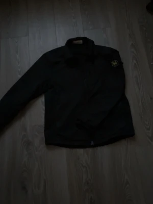 Svart jacka från Stone Island - Säljer en svart jacka från Stone Island med den klassiska logotypen på ärmen. Jackan har dragkedja framtill och en stilren, enkel design. Perfekt för dig som gillar streetwear och vill ha något som sticker ut.