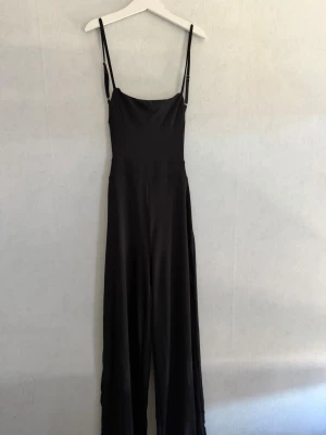 Svart jumpsuit med smala axelband - Snygg svart jumpsuit med smala, justerbara axelband och vida ben. Tillverkad i ett mjukt, lätt tyg som ger ett elegant fall. Perfekt för dig som gillar minimalistisk och stilren look. Dragkedja i sidan för enkel påklädning.