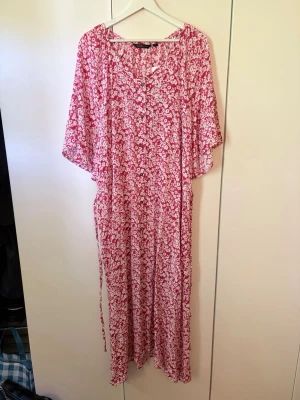 Röd & Vit blommig långklänning från Mywear Woman - Långklänning från Mywear Woman i rött & vitt blommigt mönster. Klänningen ser rosa ut men är mer röd i verkligheten. Klänningen har v-ringning, knappar framtill ända ner, korta fladdriga ärmar, löst sittande passform med knytband i midjan så det är lätt att justera passformen. Tillverkad i mjuk & luftig viskos, perfekt för varma dagar. Superfin sommarklänning! Köptes för någon månad sedan. Aldrig använd. Nypris 279kr.