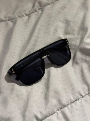 Svarta Louis Vuitton solglasögon - Snygga svarta solglasögon från Louis Vuitton med bred båge och mörka glas. Modellen har en modern, kantig form och diskreta silvriga detaljer vid fronten. Perfekta för dig som vill ha en stilren och exklusiv look.