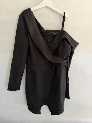 Svart oneshoulder kort klänning - Snygg svart kort klänning med unik oneshoulder-design och lång ärm på ena sidan. Klänningen har en asymmetrisk skärning och markerad midja