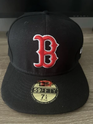 Svart Boston Red Sox keps New Era - Säljer en svart 59FIFTY keps från New Era med Boston Red Sox-logga broderad i rött och vitt framtill. Klassisk flat skärm och strukturerad form, tillverkad i slitstarkt material. Perfekt för dig som gillar sport och streetstyle.
