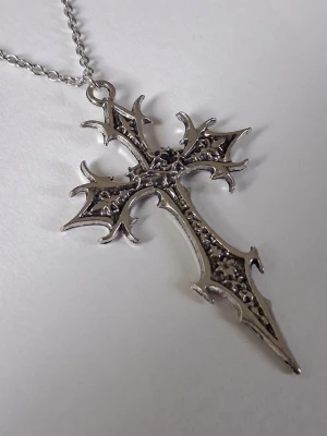 Silvrigt halsband med korshänge - Snyggt halsband med ett stort, detaljerat korshänge i silverfärgad metall. Korset har en gotisk design med spetsiga kanter och dekorativa mönster. Kedjan är tunn och matchar hängets färg perfekt. Perfekt accessoar för dig som gillar alternativ stil. Materialet rostfritt stål. 