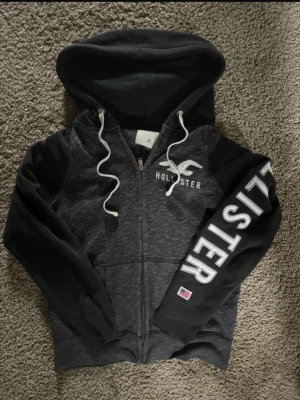 Mörkgrå Hollister hoodie med dragkedja vintage - Snygg mörkgrå hoodie från Hollister med vit logga på bröstet och stort tryck på ena ärmen. Hoodien har huva med vita snören och dragkedja framtill. Perfekt för en avslappnad och cool stil.