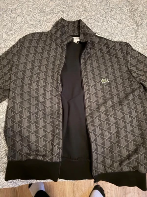 Mönstrad svart bomberjacka från Lacoste - Svart Lacoste Hoodie. Ej använd ofta säljs pga den är för liten för mig nu. Går ner i pris vid snabb affär