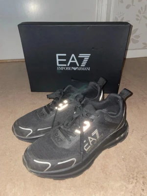 Svarta EA7 Emporio Armani sneakers - Snygga svarta sneakers från EA7 Emporio Armani med vita detaljer och logga på sidan. Skorna har en chunky sula och är tillverkade i mesh och syntetmaterial för en sportig look. Perfekta för dig som gillar stilrena och trendiga sneakers.