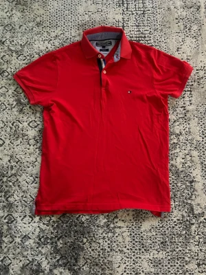 Röd pikétröja från Tommy Hilfiger - Snygg röd pikétröja från Tommy Hilfiger i slim fit-modell. Tröjan har klassisk krage med kontrastdetaljer i blått och vitt samt små knappar framtill. Liten logga på bröstet och korta ärmar. Perfekt för en stilren look.