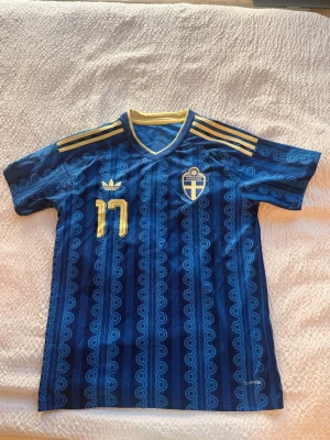 Blå Sverige fotbollströja Adidas - Säljer en blå Sverige fotbollströja från Adidas med guldiga detaljer och nummer 17. Tröjan har korta ärmar, V-ringning med gul kant och Sveriges landslagsemblem på bröstet. Mönstrad design och namnet Gyökeres på ryggen. Perfekt för match eller träning.
