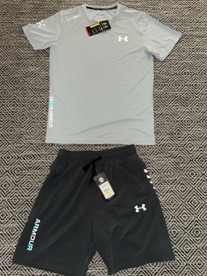 Under Armour träningsset grå - Snyggt träningsset från Under Armour med en ljusgrå t-shirt och svarta shorts. T-shirten har tryckt logga på bröstet och ärmarna, samt textdetaljer. Shortsen har dragsko i midjan och logga på benet. Perfekt för gymmet eller löpning. Storlek m passar även S
