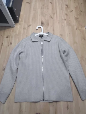 Ljusgrå ribbad ziptröja från SMDB - Ljusgrå ribbstickad tröja från SMDB med hel dragkedja framtill och klassisk krage. Tröjan har långa ärmar och en stilren, minimalistisk look som passar perfekt till många outfits. Materialet ser mjukt och bekvämt ut.