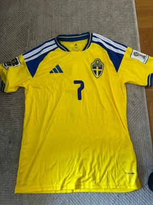 Svensk landslagströja Adidas gul - Gul svensk landslagströja från Adidas med blå detaljer och nummer 7 på bröstet. Tröjan har blå-vita ränder på axlarna, broderad landslagssköld och Adidas-logga. Tillverkad i funktionsmaterial som andas, perfekt för fotboll eller träning.