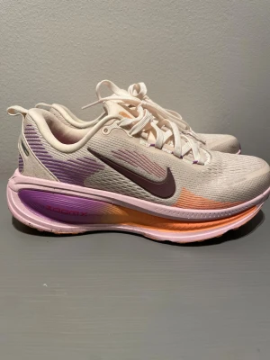 Nike ZoomX sneakers i lila och orange - Nike ZoomX sneakers i vitt med lila och orange detaljer. Skorna har en chunky sula med gradient från lila till orange, meshmaterial för bra andningsförmåga och klassisk Nike Swoosh i mörklila på sidan. Lila och röda accenter på hälen och ZoomX-logga på sulan.