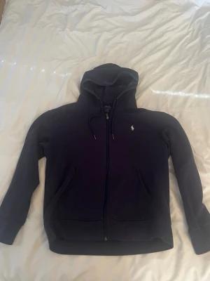 Mörkblå ziphoddie Ralph Lauren - Säljer en mörkblå ziphoodie från Polo Ralph Lauren. Klassisk design med huva och två fickor framtill. Ikoniska Polo-loggan broderad på bröstet. Perfekt för en avslappnad och stilren look. Tillverkad i mjukt bomullsmaterial. Skriv vid frågor. 