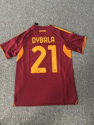 AS Roma matchtröja Dybala 21 - Snygg vinröd AS Roma matchtröja från Adidas med gula detaljer och tryck. På ryggen står det Dybala och nummer 21. Tröjan har klubbmärke och Adidas-logga på bröstet samt gula ränder på axlarna. Tillverkad i lätt och ventilerande material.