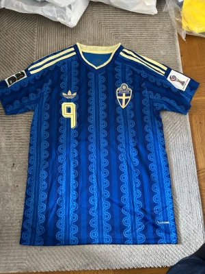 Blå Sverige fotbollströja Adidas - Retro blå fotbollströja från Adidas med Sveriges landslagsemblem och nummer 9 på bröstet. Tröjan har gula detaljer, klassiska Adidas-ränder på axlarna och ett unikt mönster i tyget. Perfekt för dig som älskar fotboll och vill sticka ut på planen.