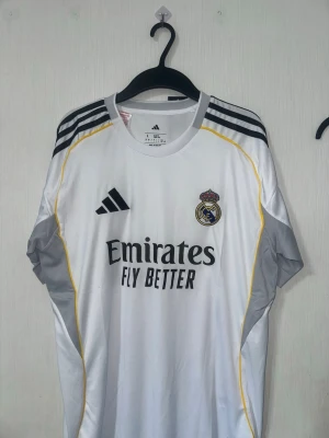 Real Madrid matchtröja Adidas vit - Snygg vit Real Madrid matchtröja från Adidas med svarta detaljer och gula linjer. Tröjan har klubbmärke på bröstet, sponsorlogga 'Emirates Fly Better' och HP-logga på ärmen. Tillverkad i lätt och ventilerande material, perfekt för fotboll eller träning.