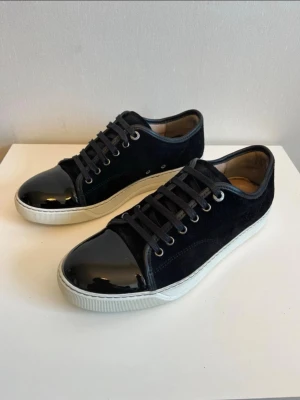 Svarta lanvins  - Snygga svarta sneakers med ovandel i mocka och en glansig, lackad tådel. Skorna har vita sulor och svarta skosnören. Metallfärgade öljetter ger en extra detalj. Perfekta för dig som gillar en stilren men ändå unik look.