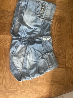 Ljusblå denimshorts  - Säljer ett par ljusblå denimshorts från G&G med dubbla knappar fram, dekorativ detalj på fickan och klassiska bakfickor. Shortsen har låg midja och är perfekta för varma dagar. Snygg tvättad look och sköna att bära.