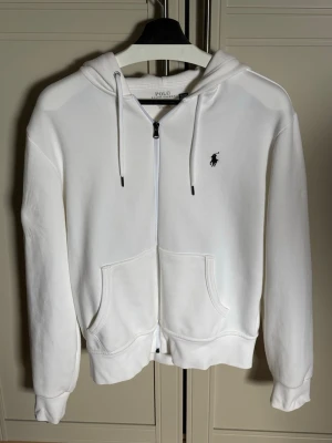 Vit hoodie från Polo Ralph Lauren. Det är en liten defekt som man ser på sista sista sidan men som knappt syns alls då den är på undersidan av armen. - Vit hoodie med dragkedja från Polo Ralph Lauren. .