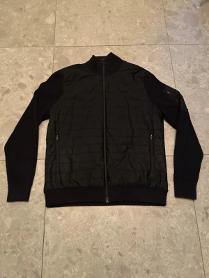 J.lindeberg cardigan - Snygg svart cardigan med quiltad framsida och ribbade muddar. Jackan har dragkedja framtill och två sidofickor med dragkedja. Samt en innerficka. Ärmarna är stickade vilket ger en schysst kontrast till den quiltade kroppen. Ytterligare en j.lindeberg logga finns på höger arm. Skriv gärna vid funderingar. Skickar gärna fler bilder. Ny pris 3200!