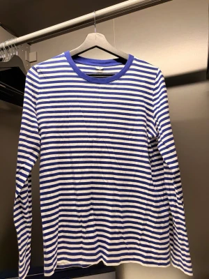 Randig långärmad t-shirt  - Säljer en långärmad t-shirt med blå och vita horisontella ränder. Tröjan har rund halsringning med blå kant och är gjord i mjuk bomull. Perfekt för en avslappnad och stilren look.