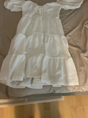 Vit långklänning med puffärm, använd en gång  - Romantisk vit långklänning med puffärmar och snörning framtill. Klänningen har flera volanglager och är tillverkad i ett lätt, luftigt tyg som passar perfekt för varma dagar. Söt och trendig modell med omlottkänsla och markerad midja.