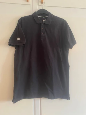 Svart pikétröja från Helly Hansen - Klassisk svart pikétröja från Helly Hansen med korta ärmar och knappslå framtill. Tröjan har HH-logga broderad på ärmen och vid kragen. Tillverkad i mjuk bomull som känns skön mot huden. Perfekt till jeans eller shorts.