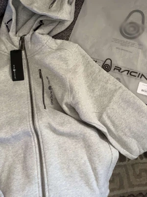 Ljusgrå hoodie från Sail Racing - Snygg ljusgrå hoodie från Sail Racing med dragkedja framtill och logga på bröstet. Hoodien har en huva med snörning och en bröstficka med dragkedja. Tillverkad i mjukt sweatshirtmaterial som känns riktigt skönt mot huden. Ny med prislapp och köpt för 1000kr.