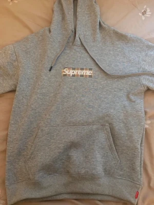 Grå Supreme hoodie med Burberry-detalj - Snygg grå hoodie från Supreme med klassisk Burberry ruta och Supreme-logga på bröstet. Hoodien har stor känguruficka, dragsko i huvan .