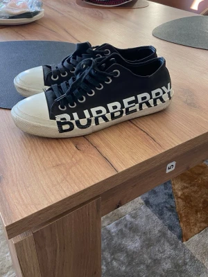 Svarta Burberry sneakers med logga - Svarta låga sneakers från Burberry med stor vit logga på sidan. Skorna har vita tåhättor och sulor, svarta skosnören och metallfärgade öljetter. Tillverkade i canvasmaterial med klassisk streetstil. Se bilder för lite skada vid hälen