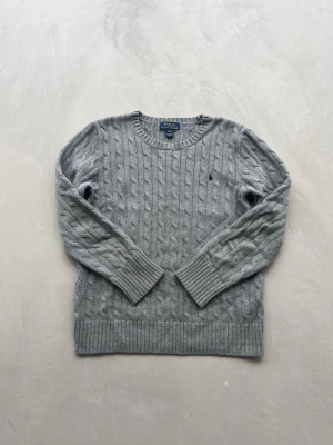 Grå kabelstickad tröja Polo Ralph Lauren - OBS❗️ALLT KOMMER STRYCKAS INNAN FRAKT! Snygg grå kabelstickad tröja från Polo Ralph Lauren med rund halsringning och diskret broderad logga på bröstet. Tröjan har långa ärmar och ribbade muddar vid ärmslut och nederkant. Perfekt för lager på lager och klassisk stil. Passar dem runt 160-170 cm lång. Inga synliga defekter