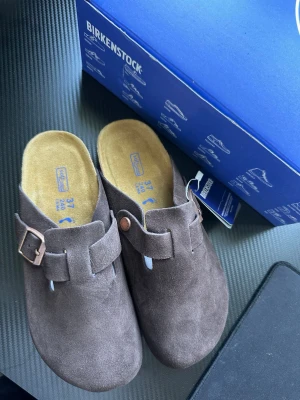 Bruna Birkenstock Boston tofflor - Säljer helt nya par klassiska Birkenstock Boston tofflor i brun mocka med justerbart spänne i metall. De har en mjuk innersula och tidlös design som passar perfekt för chill dagar hemma eller ute. Ikonisk modell som är både bekväm och stilren.