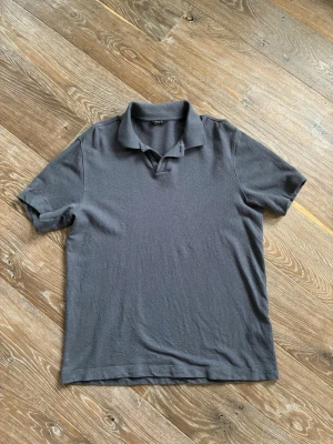 Mörkgrå pikétröja från Massimo Dutti - En stilren mörkgrå pikétröja från Massimo Dutti med klassisk krage och korta ärmar. Tröjan är tillverkad i bomull och har en enkel, tidlös design utan knappar framtill. Perfekt för en avslappnad men snygg look.
